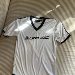 Vit tröja från Unihoc strl 140 - Snygg vit fotbollströja från Unihoc med svart V-ringad krage och svarta detaljer vid ärmslut. Tröjan har korta ärmar och är tillverkad i ett lätt och funktionellt material som passar perfekt för träning. Stor Unihoc-logga på bröstet.