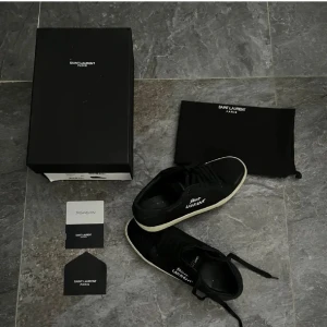 Svarta sneakers från Saint Laurent - Svarta sneakers från Saint Laurent med vit sula och vit logotyp på sidan. Klassisk låg modell med snörning och rund tå. Ovandel i mocka och detaljer i skinn. Snygg och stilren design som passar till det mesta.