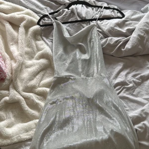 Glittrig silver klänning med snörning - Supersnygg klänning i glittrigt silvertyg med tunna axelband och öppen rygg med snörningsdetalj. Klänningen har en elegant, figurnära passform och skimrar i ljuset. Perfekt för dig som vill sticka ut och glänsa på festen!