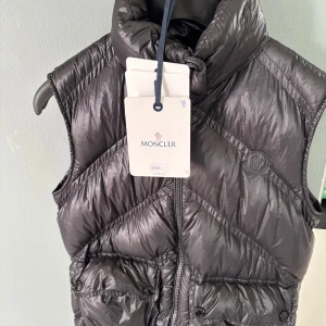 Svart dunväst från Moncler - Snygg svart dunväst från Moncler med glansig finish och quiltad design. Västen har hög krage, dragkedja framtill och två stora fickor med knappar. Ikonisk Moncler-logga på bröstet och innerficka med tygpatch. Perfekt för lager-på-lager-stil. .