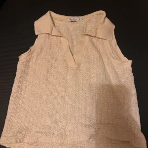 Beige ärmlös linnetopp Stradivarius - Superfin beige linnetopp från Stradivarius med krage och v-ringning. Toppen är ärmlös och har en stickad struktur som ger en cool och avslappnad vibe. Perfekt till jeans eller kjol för en fräsch look.