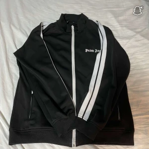 Palm Angels svart track jacket - Svart track jacket från Palm Angels med vita dubbla ränder längs axlar och ärmar. Dragkedja framtill, hög krage och fickor på sidorna. Snygg streetwear-vibe och klassisk logga på bröstet. Perfekt till sneakers och jeans.