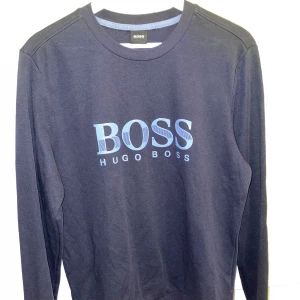 Mörkblå tröja från Hugo Boss - Snygg mörkblå långärmad tröja från Hugo Boss med stort ljusblått BOSS-tryck på bröstet. Klassisk rund halsringning och normal passform. Perfekt för dig som gillar stilrena och trendiga plagg med tydlig logga.