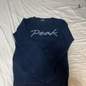 Mörkblå stickad Peak Performance tröja - Mörkblå stickad tröja från Peak Performance med broderad logga framtill. Klassisk rund halsringning och långa ärmar. Ribbstickade muddar vid ärmslut och nederkant ger en snygg passform. Perfekt för dig som gillar stilren och sportig look.