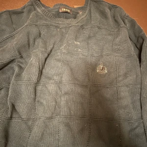 Grå stickad tröja från IZOD - Mörkgrön stickad tröja från IZOD med rutigt strukturmönster och broderad logga på bröstet. Klassisk rund halsringning och långa ärmar. Perfekt för dig som gillar en stilren och enkel look med lite preppy känsla.