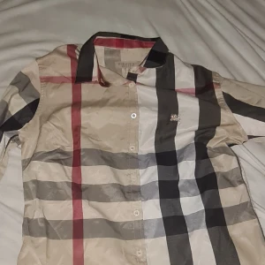  Rutig skjorta från Burberry - Snygg rutig skjorta från Burberry i klassiska färger med stora svarta, röda och beige rutor. Skjortan har knappar framtill, krage och lång ärm. Perfekt för dig som gillar stilren design med ikoniskt mönster.