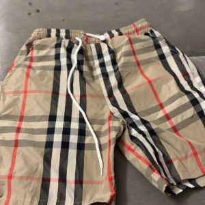 Burbery shorts - Snygga beige shorts från Burberry med klassiskt rutigt mönster i svart, vitt och rött. De har elastisk midja med vit snörning och är tillverkade i lätt polyester, perfekt för varma dagar. Passformen är normal och de har en modern streetstil.