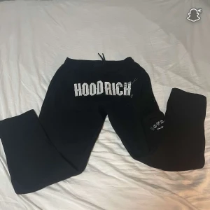 Svarta mjukisbyxor från Hoodrich - Svarta mjukisbyxor från Hoodrich med stor vit logga framtill och mindre tryck på benet med liten ficka. Byxorna har snörning i midjan och är gjorda i mjukt bomullsmaterial. Perfekta för chill eller streetwear och har en loose passform för skön vibe.