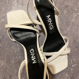 Vita sandaler med klack från MNG - Stilrena vita sandaler från MNG med smala remmar och fyrkantig tå. Skorna har svart innersula och en elegant låg klack. Perfekta för dig som gillar minimalistisk och trendig design. Materialet är syntet och de har en modern look.