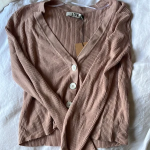 Ribbad beige kofta från JDY - Superfin ribbad kofta från JDY i en mjuk beige ton. V-ringad med stora vita knappar framtill och långärmad modell. Perfekt att slänga över en topp för en chill och snygg look. Materialet är stretchigt och känns skönt mot huden.