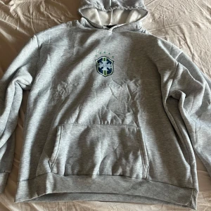 Grå hoodie med Brasilien tryck och huva - Snygg grå hoodie med tryck på bröstet i blått och grönt. Klassisk känguruficka framtill och huva med dragsko. Perfekt för chill dagar eller när du vill ha en enkel och stilren look. Kan gå ner i pris 