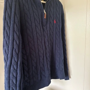 Marinblå kabelstickad tröja Polo Ralph Lauren - Kabelstickad marinblå tröja från Polo Ralph Lauren med halv dragkedja och röd broderad logga på bröstet. Tröjan har lång ärm, ribbade muddar och är tillverkad i mjuk ull. Perfekt för dig som gillar klassisk och stilren look.