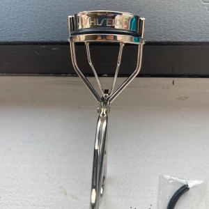 Shiseido ögonfransböjare med extra gummi - helt ny aldrig andvänd snygg och klassisk ögonfransböjare från Shiseido i silverfärgad metall. Ergonomisk design med bred böjdel och mjukt svart gummiband för skonsam böjning av fransarna. Medföljer ett extra gummiband i förpackning. Perfekt för att få snygg böj på fransarna. 
