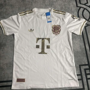 Vit Bayern München Musiala tröja - Säljer en vit Bayern München 125-års jubileumströja från Adidas med Musiala 10 på ryggen. Tröjan har guldiga detaljer, klassiska Adidas-ränder på axlarna och klubbmärke framtill. Tillverkad i lätt och ventilerande material (AEROREADY). Perfekt för dig som älskar fotboll och snygg design.
