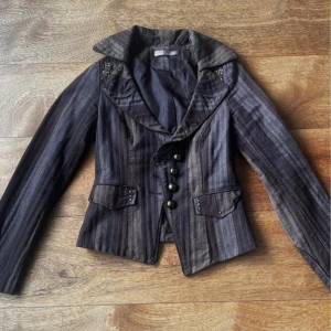 Randig kavaj från Vila i brun och grå - Snygg kavaj från Vila i storlek XS med randigt mönster i bruna och grå toner. Kavajen har markerad krage, dekorativa knappar framtill och fickor med lock. Långärmad och figurnära passform, perfekt för en cool och dressad look.