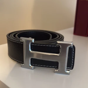 Svart skärp med H-spänne - Säljer ett svart skärp  med ett stort, silvrigt H-spänne. Bältet är gjort i skinn och har vita kontrastsömmar längs kanterna som ger en snygg detalj. Perfekt accessoar för att lyfta din outfit med en lyxig touch. Sizen är 115 men har hål mindre än vanligt 