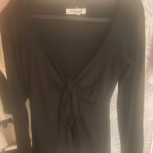 Svart långärmad klänning Design By si - Svart långärmad klänning från Design S med djup v-ringning och snygg knytning framtill. Klänningen har en smickrande passform och är perfekt för dig som gillar stilrena plagg med en twist.