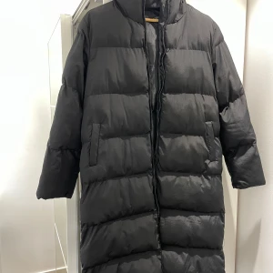 Svart lång pufferjacka med huva - Lång svart pufferjacka med huva och dragkedja framtill. Jackan har en quiltad design med breda kanaler och två sidofickor. Perfekt för kalla dagar när du vill hålla dig varm och stilsäker.