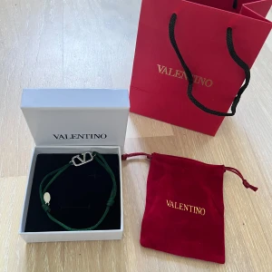 Grönt armband från Valentino - Snyggt armband från Valentino med grönt band och silverfärgat V-logo i metall. Justerbar knytning och exklusiv känsla, perfekt för dig som gillar stilrena accessoarer. Kommer med originalask och röd sammetspåse med guldfärgad logga.