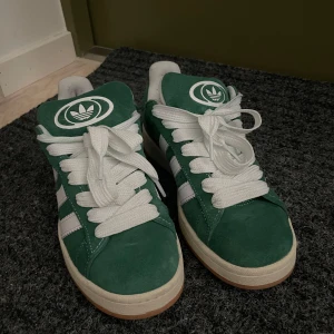 Adidas Campus gröna - Gröna Adidas Campus sneakers i mocka med vita detaljer och klassiska tre ränder på sidan. Skorna har vit sula, rund tå och snörning. Extra gröna skosnören medföljer. Perfekta för dig som vill sticka ut med en retro vibe.