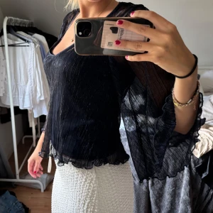 Svart glittrig blus med vida ärmar - Supersnygg svart blus med glittriga detaljer och transparenta, vida ärmar. Blusen har v-ringning och volangkant nertill och vid ärmslut. Perfekt för dig som vill sticka ut lite extra med en elegant och trendig look.