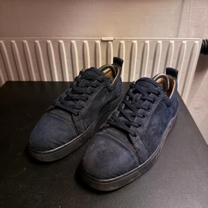 Mörkblå mockasneakers från Louboutin - Snygga mörkblå sneakers i mocka från Christian Louboutin med klassisk röd sula och broderad logga på hälen. Skorna har rund tå, snörning och en stilren design som sticker ut med exklusiva detaljer och lyxig känsla. Passar även 42, har använts en del och har slitage under sulan och i hälen.