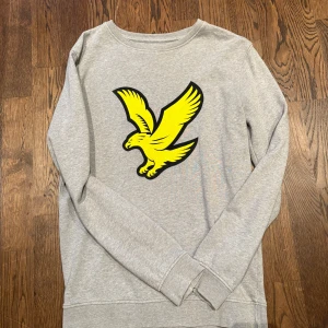 Grå sweatshirt från Lyle & Scott - Snygg grå sweatshirt från Lyle & Scott med stort gult fågeltryck på bröstet. Tröjan har rund hals och långa ärmar, perfekt för en chill och stilren look. Tillverkad i mjuk bomull som är skön att ha på sig hela dagen. Pris kan diskuteras vid snabb affär 😁
