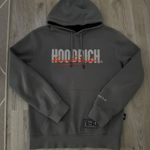 Grå hoodie från Hoodrich med tryck - Snygg grå hoodie från Hoodrich med stor logga i vitt och rött framtill och på ryggen. Tröjan har huva med snörning, magficka och ribbade muddar. Perfekt för en chill streetwear-look. Detaljerad patch nertill och broderad text på ärmen.