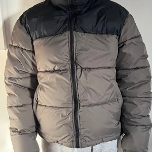 Grå och svart pufferjacka från Everest - Säljer en snygg pufferjacka från Everest i grått och svart. Jackan har hög krage, dragkedja framtill och breda, vadderade paneler som ger en cool och sportig look. Perfekt för kalla dagar när du vill hålla värmen och ändå se stilren ut.