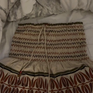 Beige kjol med mönster och snörning - Supersöt beige kjol med elastisk smock i midjan och snörning framtill. Kjolen har ett unikt mönster i brunt och grönt nertill och är perfekt för dig som gillar boho-stil. Lätt och luftig, passar perfekt till varmare dagar. lite lösa trådar vid kjolen men inget man tänker på. står ej storlek men skulle tippa på Xs.