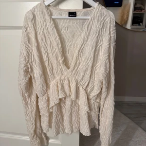Krämvit blus med volang från Gina Tricot - Supersöt krämvit blus från Gina Tricot med v-ringning och volangdetalj framtill. Blusen har långärmade ärmar och ett strukturerat, lite skrynkligt tyg som ger en cool look. Perfekt att styla med jeans eller kjol för en trendig outfit.