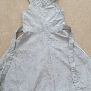 Klänning - Supersöt ljusgrå klänning från Lindex med broderade detaljer och små paljetter i midjan. Klänningen har omlottband i sidan och dragkedja bak. Perfekt för dig som gillar en enkel men ändå glittrig look. Har en fläck och bandet bakom klänningen har gått av.