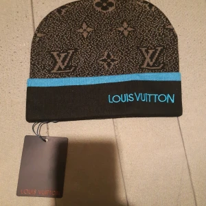 Louis Vuitton svart mössa med blå kant - Snygg svart mössa från Louis Vuitton med klassiskt monogrammönster i grått och en blå kant. Broderad Louis Vuitton-logga i blått framtill. Perfekt accessoar för dig som vill sticka ut med lyxig streetstyle. Materialet känns mjukt och stretchigt.