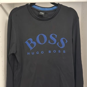 Svart långärmad tröja Hugo Boss - Svart långärmad tröja från Hugo Boss med stort blått BOSS-tryck på bröstet. Klassisk rund halsringning och enkel design utan extra detaljer. Perfekt för dig som gillar stilrena och snygga logoplagg.