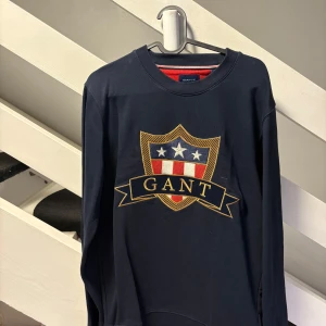 Mörkblå GANT sweatshirt med brodyr - Mörkblå sweatshirt från GANT med stort broderat sköldmärke framtill i rött, vitt och guld. Klassisk rund halsringning med ribbade muddar och insida i rött. Perfekt för dig som gillar preppy stil och tydliga loggor.