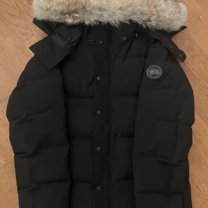 Svart Wyndham jacka Canada Goose - Säljer en svart dunjacka från Canada Goose  jacka snygg fuskpäls runt kanten. Jackan har tydliga paneler, knappar framtill och dragkedja. Klassisk patch på ärmen och två fickor på sidorna. Perfekt för kalla dagar. Skriv för mer bilder.