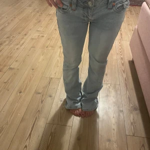 Ljusblå True Religion bootcut jeans - Säljer ett par ljusblå bootcut lågmidjade jeans från True Religion med ikoniska bakfickor och orange broderi. Jeansen har låg midja, klassisk femficksdesign och är gjorda i mjukt denim. Perfekta för dig som gillar en avslappnad och trendig look.