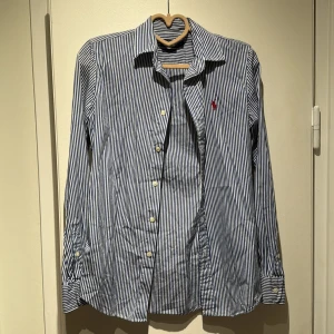 Randig skjorta från Polo Ralph Lauren - Snygg blå- och vitrandig skjorta från Polo Ralph Lauren med klassisk krage och lång ärm. Custom fit-modell med knappar framtill och ikonisk röd logga broderad på bröstet. Perfekt för dig som gillar stilrena och tidlösa plagg.