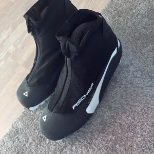 Svarta Fischer pjäxor strl 41 - Säljer ett par svarta Fischer pjäxor med Comfort Guard insulation. De har dragkedja framtill, snörning under locket och detaljer i vitt. Perfekta för längdskidåkning och håller fötterna varma under vintern.