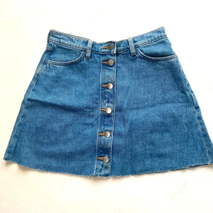 Blå jeanskjol med knappar framtill - Snygg blå jeanskjol i A-linjeform med fem metallknappar framtill, rå kant nertill och klassiska fickor både fram och bak. Perfekt för dig som gillar en avslappnad men trendig stil. Passar till allt från sneakers till boots. Storlek känns mer som s