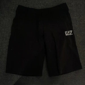 Svarta EA7 Emporio Armani shorts - Svarta shorts från EA7 Emporio Armani med vit logga på vänster ben. Klassisk rak passform och elastisk midja för skön känsla. Perfekta för chill eller sport, med stilren design och mjukt material.