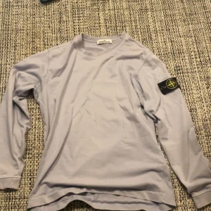Ljusgrå långärmad Stone Island tröja - Snygg ljusgrå långärmad tröja från Stone Island med klassisk patch på vänster ärm. Tröjan har rund halsringning och är gjord i mjuk bomull, perfekt för en clean och stilren look. Passar dig som gillar streetwear och vill ha något enkelt men ändå fett.