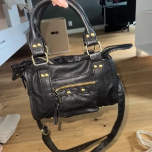 Svart axelväska från Misako i skinn - Säljer en svart axelväska från Misako i skinnimitation med guldfärgade detaljer och dragkedja framtill. Väskan har både handtag och justerbar axelrem, perfekt för att bära på olika sätt. Snygg och stilren modell med coola nitar och praktiska fack. Väldigt lik en city bag