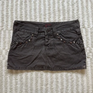 Mörkgrå jeanskjol med färgglada detaljer - Snygg grå kort jeanskjol från Miss Sixty med coola nitar och dragkedjor på bakfickorna. Fickorna har färgglada broderier och kjolen har bälteshällor och knappstängning. Så snyggt med unika detaljer!!