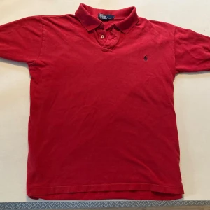 Röd pikétröja från Polo Ralph Lauren - Klassisk röd pikétröja från Polo Ralph Lauren med broderad logga på bröstet. Tröjan har krage, knäppning med två knappar och är kortärmad. Tillverkad i bomull för en skön och avslappnad känsla. Fint skick