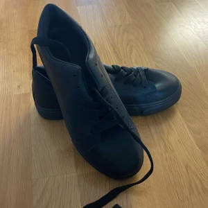 Svarta sneakers med snörning - Stilrena svarta sneakers med platt sula och rund tå. Skorna har klassisk snörning och är tillverkade i ett syntetmaterial som ger en clean look. Perfekta för dig som gillar minimalistisk stil och vill ha ett par skor som funkar till det mesta.