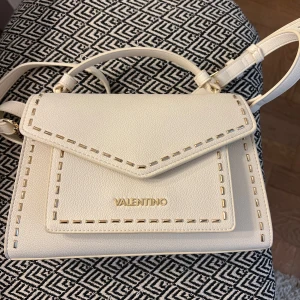 Vit axelväska från Valentino i skinn - Snygg axelväska från Valentino i vit färg med gulddetaljer och dekorativa sömmar. Väskan är tillverkad i skinn och har ett stilrent, fyrkantigt format med justerbar axelrem. Perfekt för dig som vill ha en clean och lyxig look.