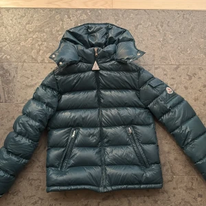 Mörkgrön dunjacka från Moncler - Säljer en snygg mörkgrön dunjacka från Moncler med glansig finish och huva. Jackan har dragkedja framtill, två fickor med dragkedja och klassisk Moncler-logga på ärmen. Perfekt för kalla dagar och riktigt skön att bära. den är knappt använd, köptes på monclers hemsida för 9550 köptes 2024. skriv för fler bilder eller frågor💕