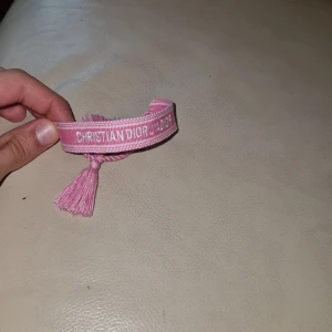 Rosa armband från Christian Dior - Säljer ett snyggt rosa armband från Christian Dior med broderad logga och tofsar i ändarna. Armbandet är gjort i textil och har en cool, avslappnad vibe som passar perfekt till din outfit. Justerbar storlek med knytning.