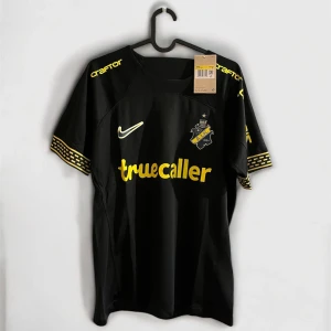 AIK svart matchtröja Nike 2024 - AIK:s officiella svarta matchtröja från Nike med gula detaljer och tryck. Kortärmad modell med gul logga, sponsortryck och mönstrade ärmslut. Materialet är lätt och andas, perfekt för träning eller match. Snygg design med klubbmärke på bröstet och tryck på ryggen.